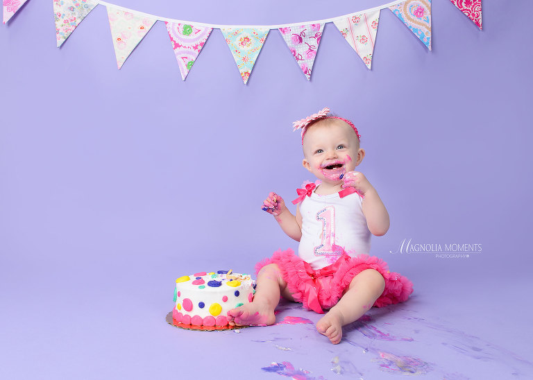 Adorable baby girl dressed in a cute cake smash outfit on a purple background with her smash cake. Take by Evan Pollock of Magnolia Moments Photography, who does Cake Smash photography and 1st birthday photography near me in Collegeville PA.
