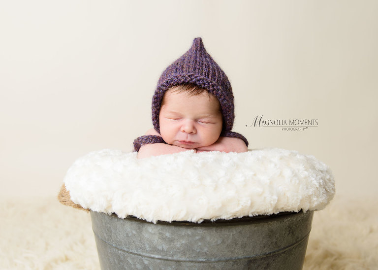 Newborn girl posed in bucket wearing purple hat as her newborn baby clothes in her newborn baby pic by Evan Pollock one of the professional photographers near me. 