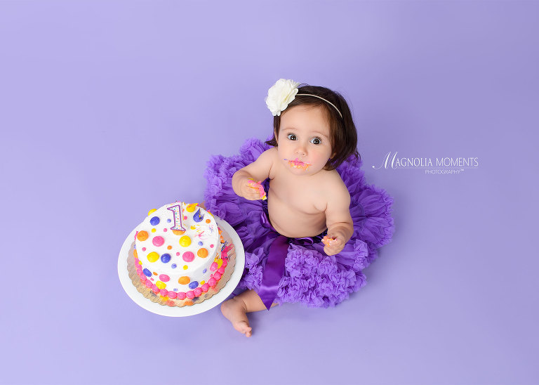 Adorable baby girl in purple tutu with birthday cake during her first birthday cake smash photography session by Magnolia Moments Photography