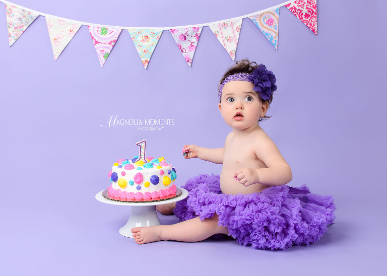 Precious baby girl in purple tutu photographed on purple background with cake during her Cake Smash session by Evan Pollock of Magnolia Moments Photography a photography studio near me in PA