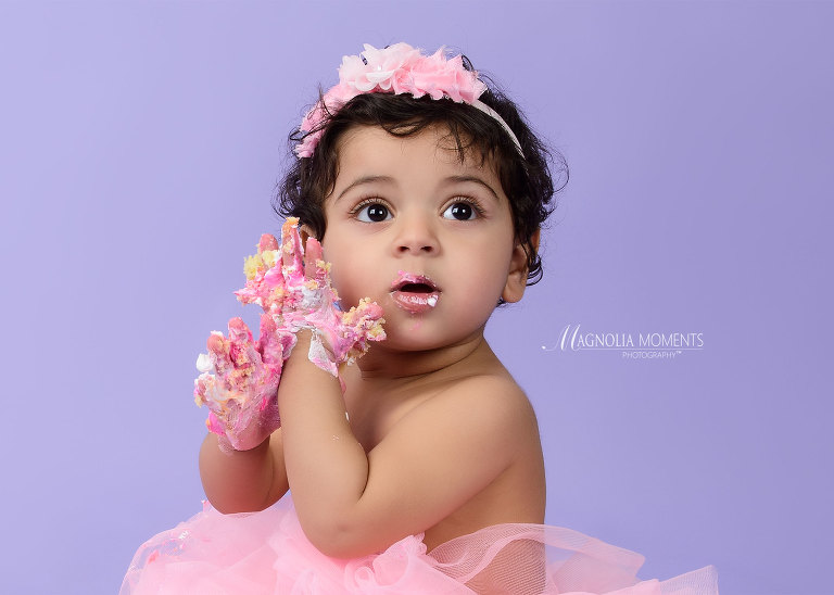close-up of baby girl in pink on lavender backdrop with hands covered in cake during her 1st birthday cake smash at Magnolia Moments Photography who does professional photography near me. Lansdale child photographer