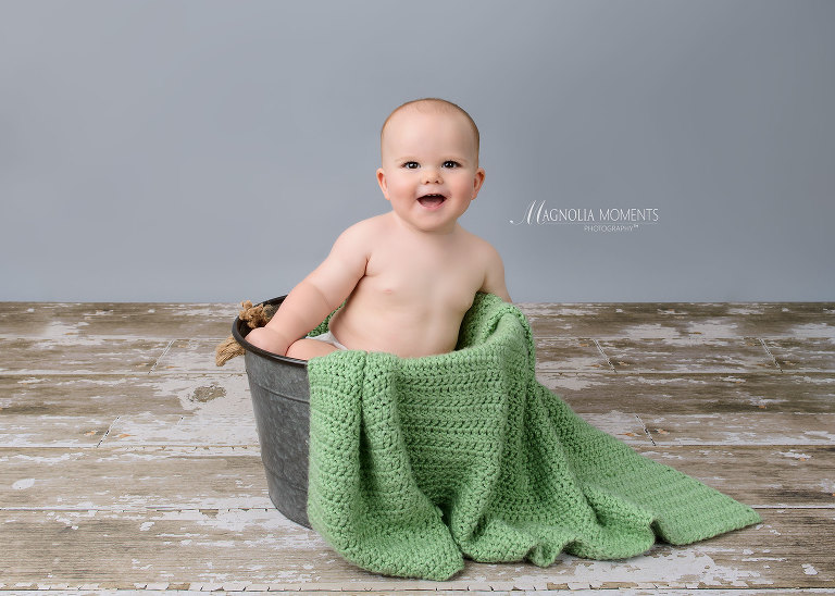 Happy baby boy in metal bucket on barn floor with green blanket during his 1st birthday photography session by Evan Pollock of Magnolia Moments Photography one of the photographers near me. Philadelphia baby photographer