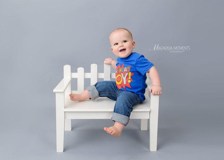 Smiling baby boy on white bench taken during his 1st year portrait session by Evan Pollock of Magnolia Moments Photography a baby and child photographer near me. Philadelphia baby photographer