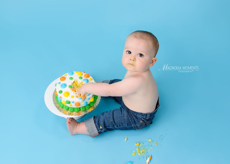 Baby boy in jeans on blue background with birthday cake during his cake smash session by Evan Pollock of Magnolia Moments Photography who does baby photography near me. Philadelphia baby photographer