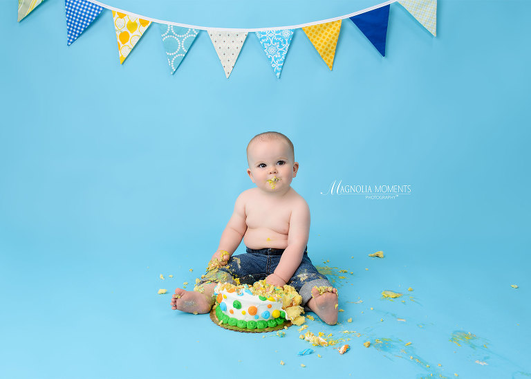 Baby boy in jeans on blue backdrop with birthday cake in his 1st birthday cake smash session by Evan Pollock of Magnolia Moments Photography who does professional photography near me. Philadelphia baby photographer