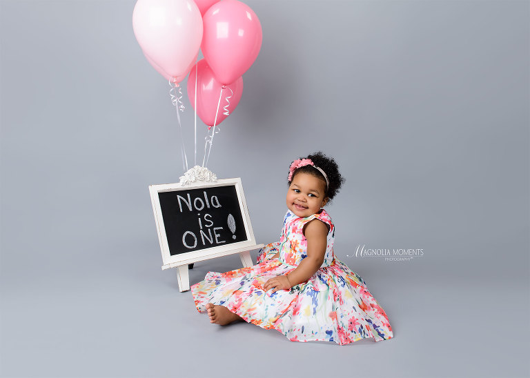 Baby girl in floral dress on grey with pink balloons during her 1st birthday cake smash and portrait session photographed by Evan Pollock of Magnolia Moments Photography as professional photographer near me.