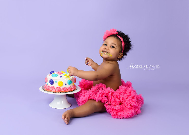 Baby girl in pink tutu as her cake smash outfit on lavender backdrop with birthday cake during her 1st birthday portrait session and cake smash by Evan Pollock of Magnolia Moments Photography who offers cake smash photography near me.