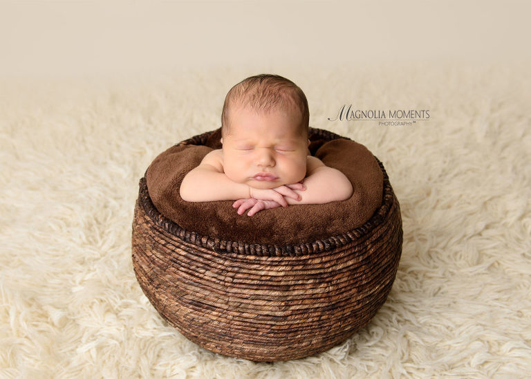 Tiny baby boy posed in brown basket with his chin on his hands for his newborn photoshoot by Magnolia Moments Photography one of the professional photography studios near me.